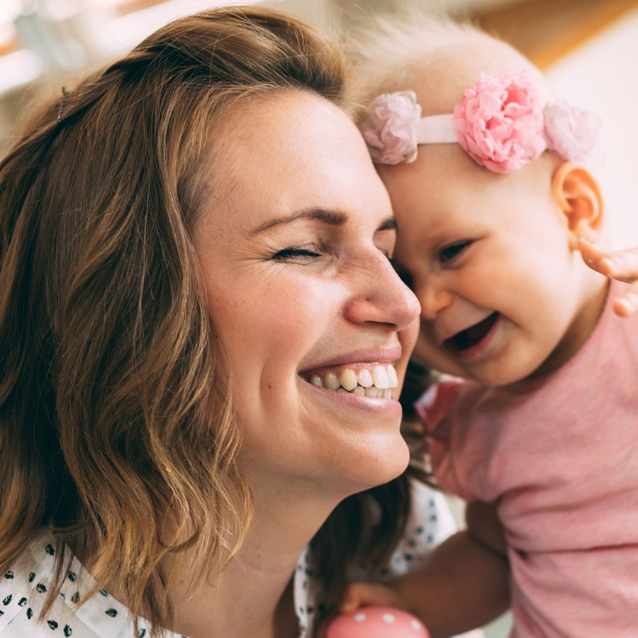 A smiling woman hugs a laughing baby wearing a pink headband and shirt. They are close together, sharing a joyful moment.