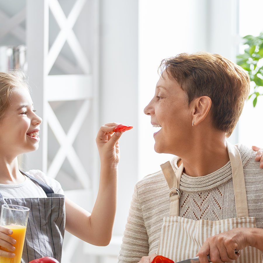 A smiling young girl offers a piece of red bell pepper to an older woman in an apron, while holding a glass of orange juice in a bright kitchen.