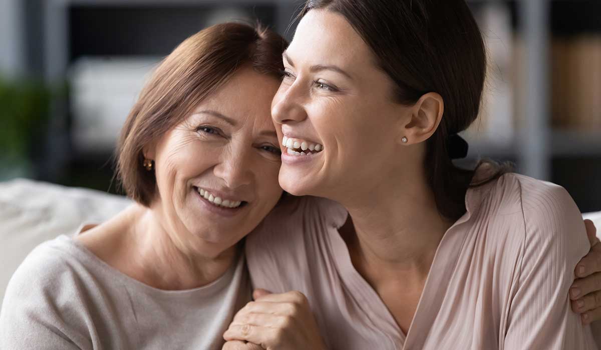 Two women sit close together on a couch, smiling and hugging. One appears older with short brown hair, and the other is younger with long brown hair, showing a warm, joyful moment.