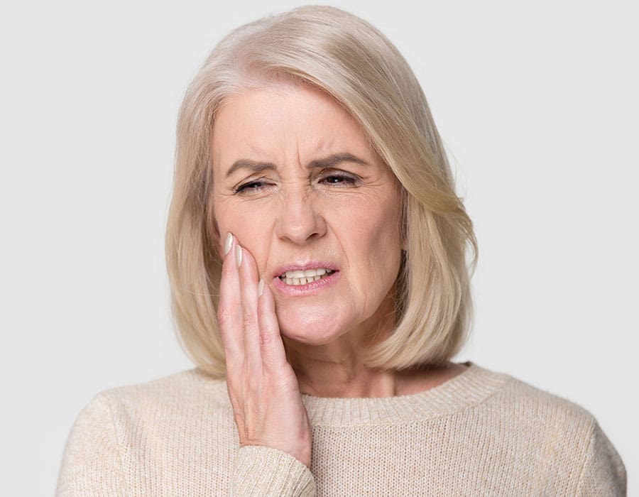 A woman sits indoors with her eyes closed, holding her cheek and wincing in pain, suggesting she has a toothache or facial discomfort.