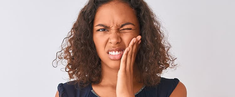 A young woman with curly hair frowns and squints, holding her hand to her cheek as if experiencing tooth or jaw pain, against a plain light background.