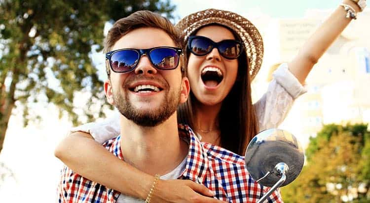 A cheerful man and woman wearing sunglasses smile and pose together outdoors, with the woman hugging the man from behind. They look happy and carefree, with bright sunlight and greenery in the background.